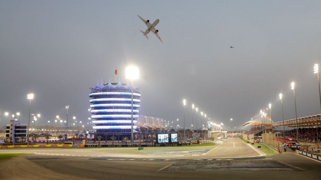 Bahrain International Circuit i kvällsbelysning, med ett flygplan som passerar över banan och huvudtornet upplyst i bakgrunden.