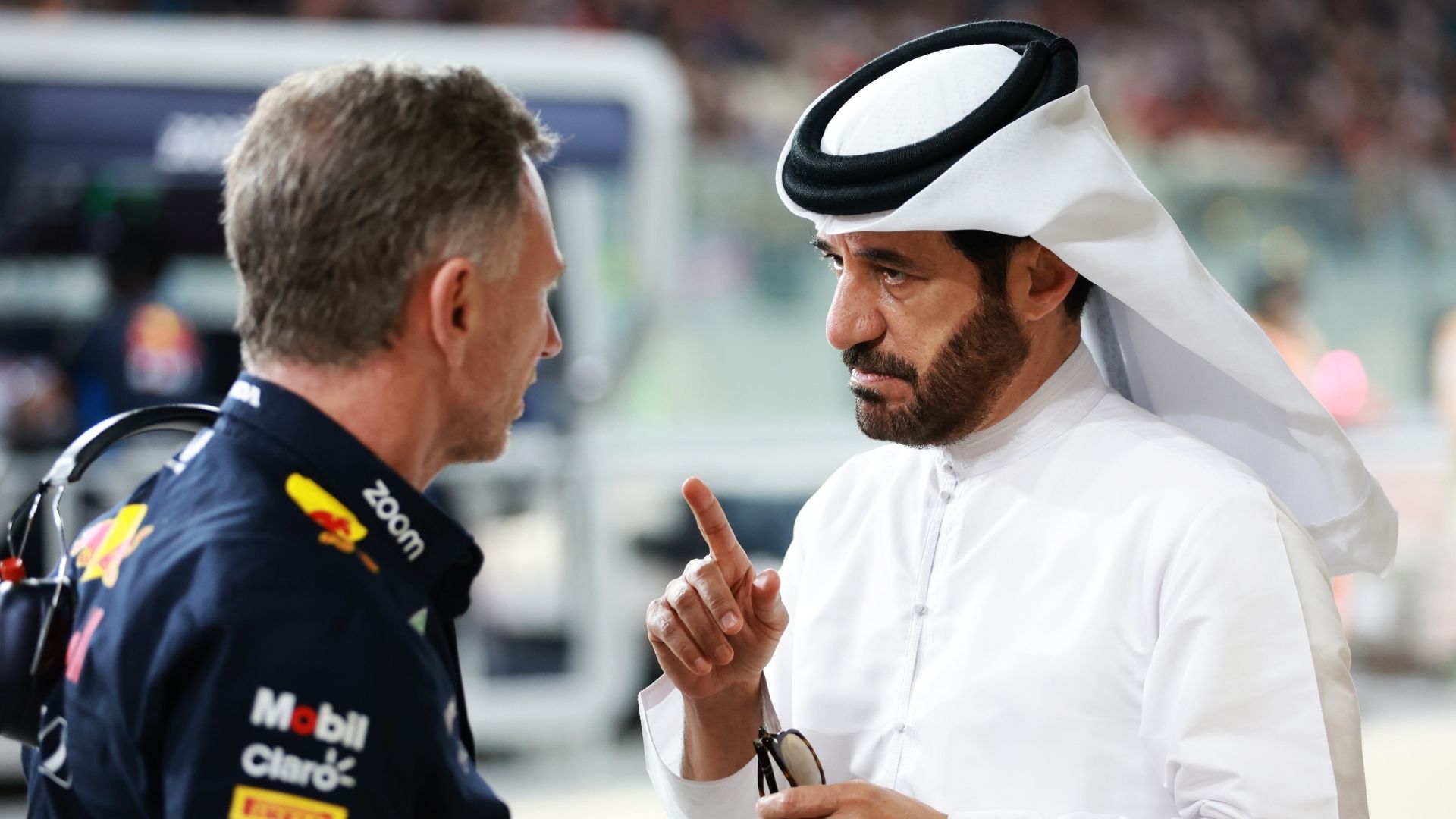 En man i Red Bull-kläder samtalar med en annan man klädd i en traditionell vit arabisk dräkt, som pekar med fingret i en intensiv diskussion i en Formel 1-depåmiljö.