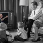 En retrobild av en familj som sitter i vardagsrummet och tittar på en Formel 1-sändning på en gammal TV, där Lewis Hamilton gör tummen upp.
