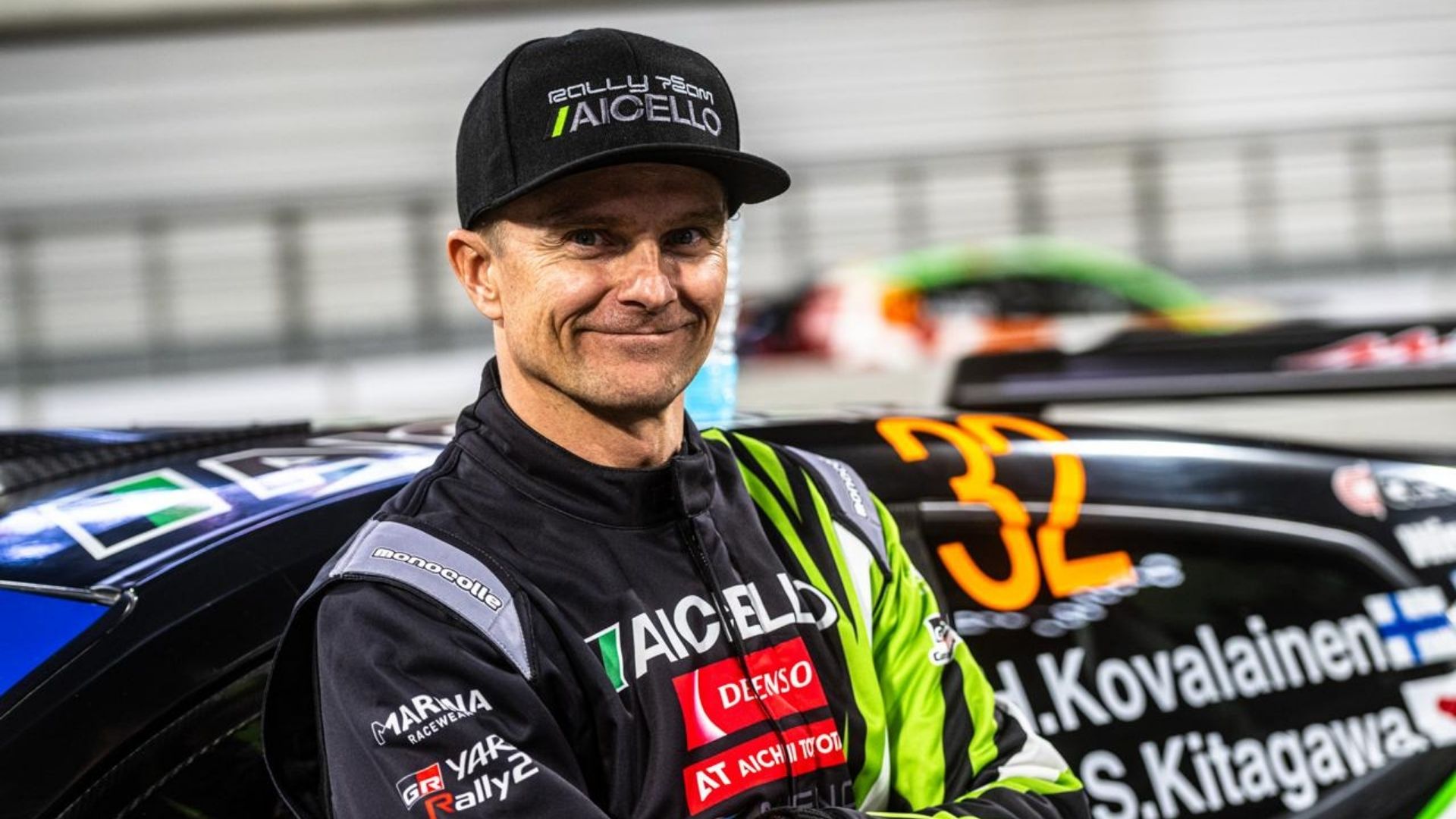 Heikki Kovalainen, klädd i sin rallyoverall från Aicello Racing, står leende framför sin bil, redo att återvända till tävling ett år efter en hjärtoperation.
