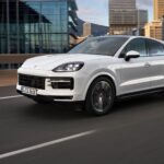 En vit Porsche Cayenne kör på en motorväg, omgiven av moderna byggnader i bakgrunden.